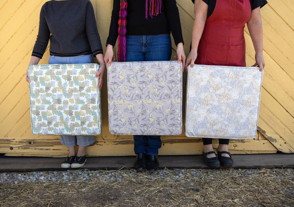 Three women standing in front of a yellow barn door holding pillows printed with the Swallowtail Farm collection.