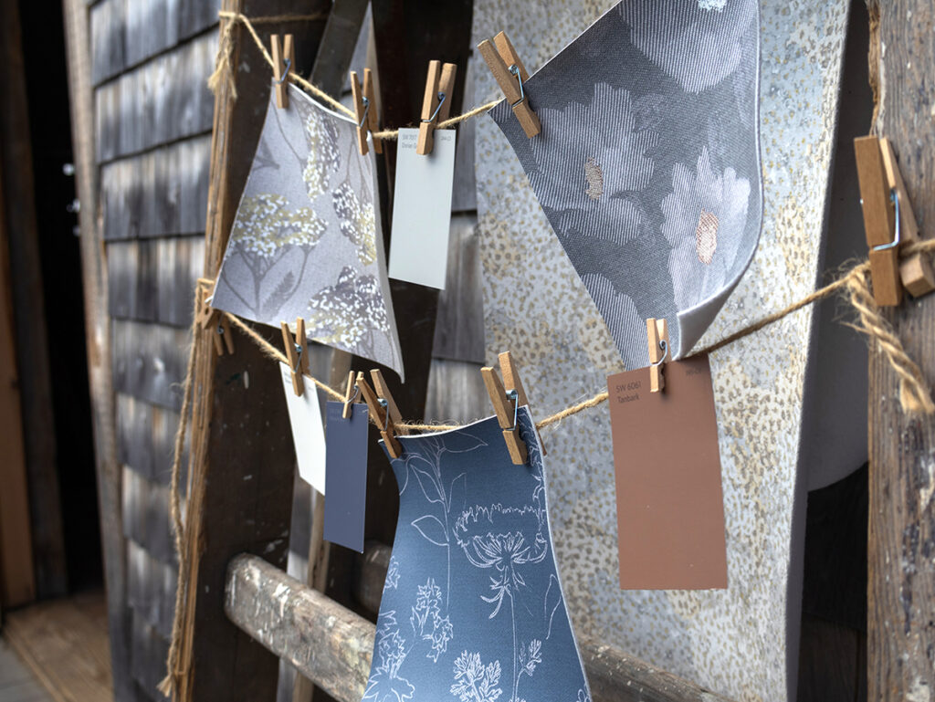 A photo of a wooden ladder leaning against a weathered barn with twine and clothespins holding swatches of Design Pool patterns from the Swallowtail Farm collection and paint chips.